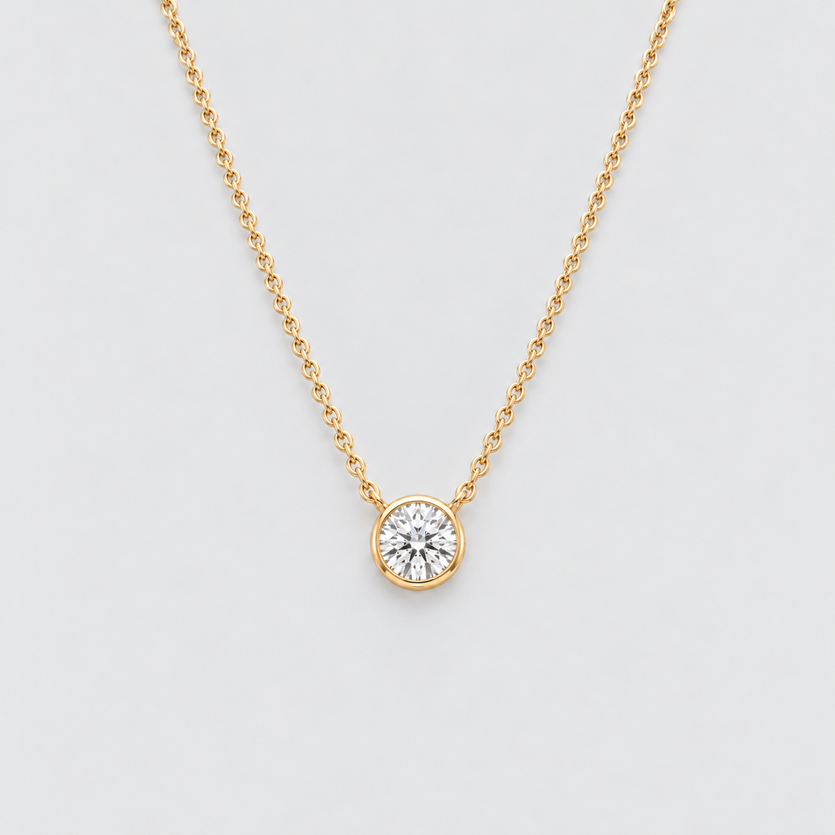 A delicate yellow gold chain necklace with a single round bezel-set diamond pendant, displayed against a clean light gray background.