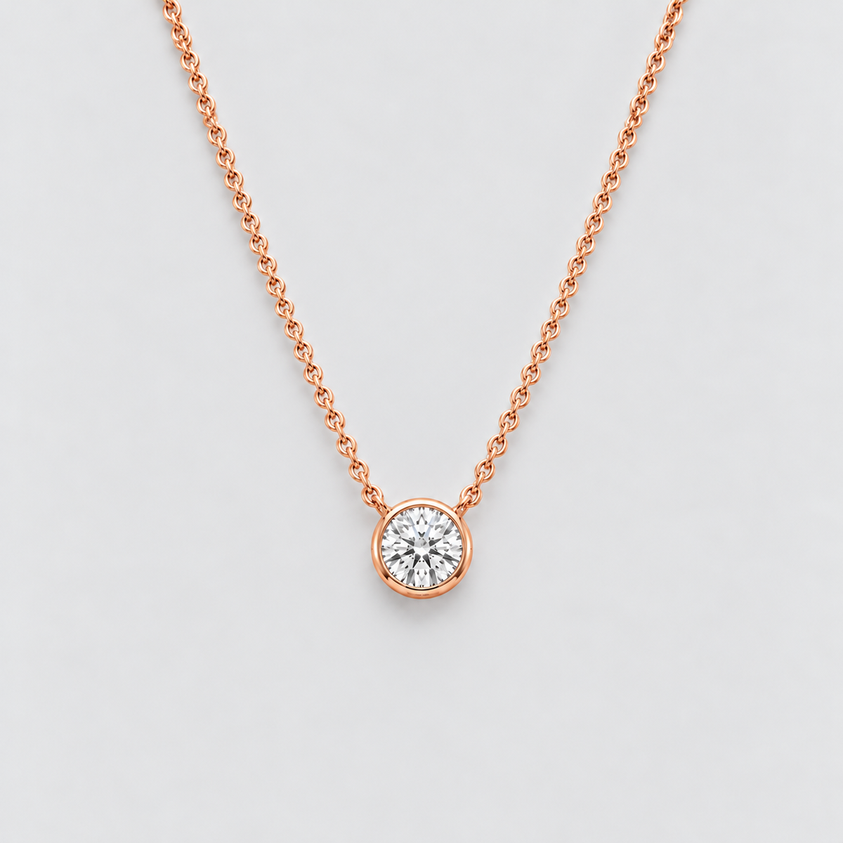 A delicate rose gold chain necklace with a single round bezel-set diamond pendant, displayed against a clean light gray background.