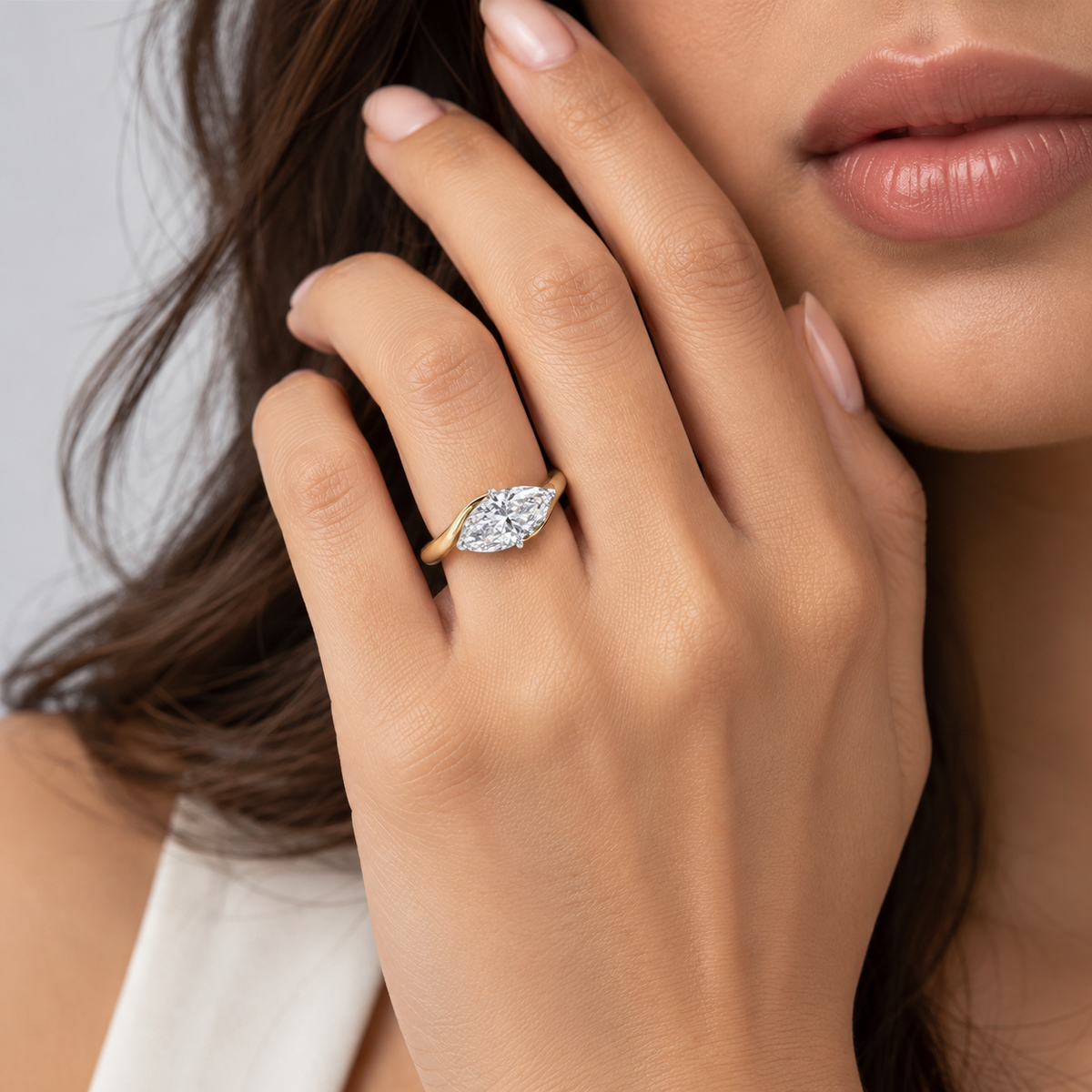 A close-up lifestyle shot of a woman wearing a yellow gold ring with a marquise-cut diamond on her finger. The ring is shown on her hand, which is resting near her face, against a soft gray background.