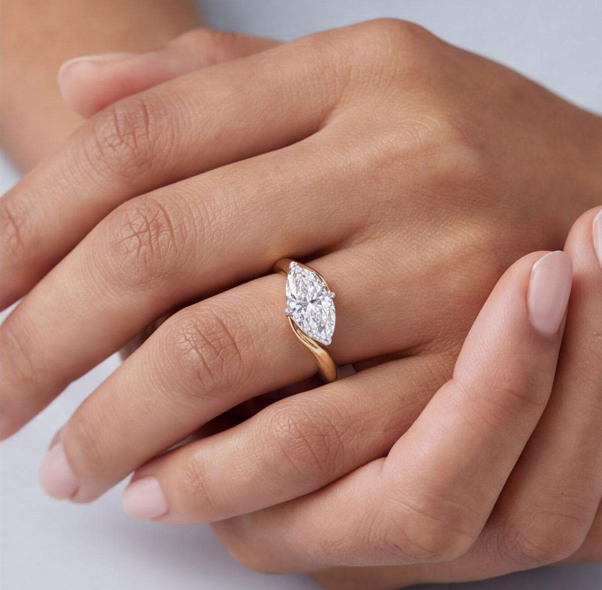 A close-up of a hand wearing a yellow gold ring with a marquise-cut diamond. The diamond is set in a curved band, and the hand is shown against a soft, light gray background.