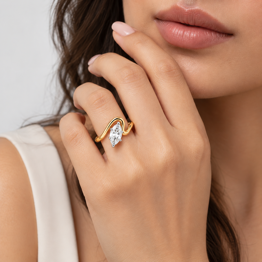 A close-up lifestyle shot of a woman's hand wearing a gold ring with a marquise-cut clear gemstone. The ring features a wavy gold band and is set against a neutral light gray background.