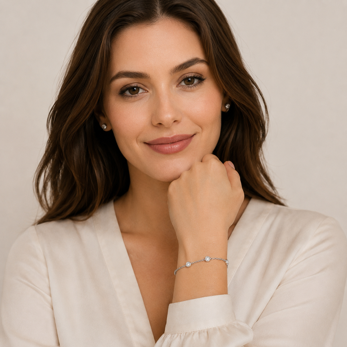 A woman with wavy brown hair wearing a delicate silver bracelet with small clear gemstones, posing in a white blouse against a soft beige background.