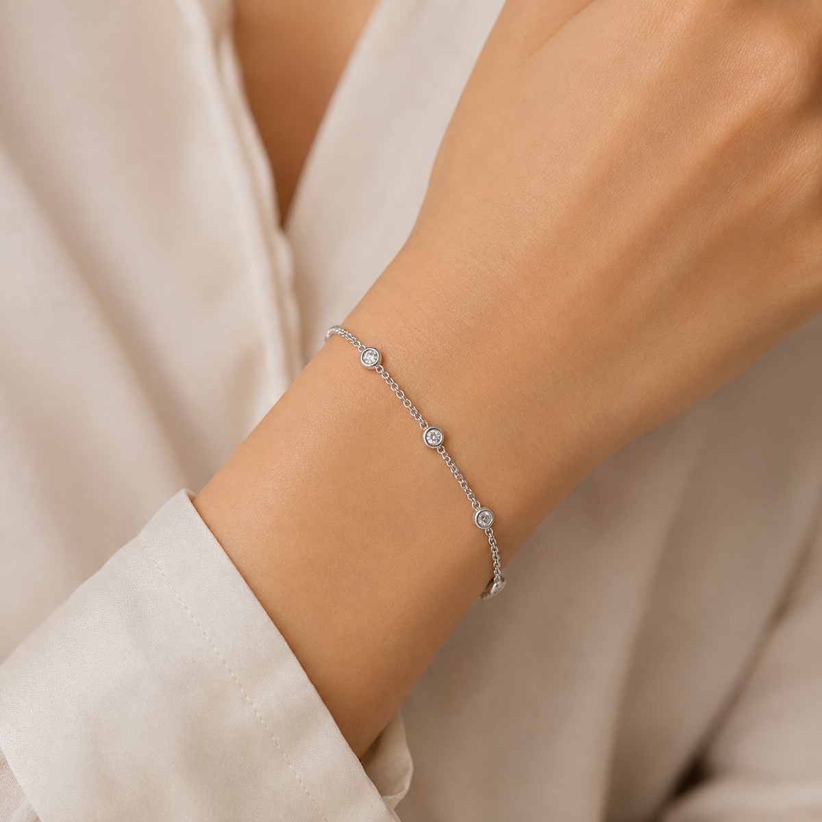 A delicate silver chain bracelet with three small round bezel-set clear stones, shown worn on a person's wrist. The person is wearing a soft, light beige blouse.