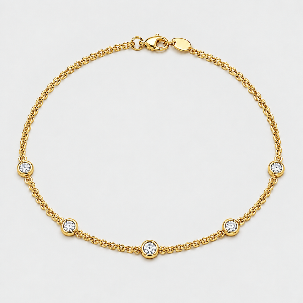A delicate gold chain anklet featuring five evenly spaced round bezel-set clear crystals on a light gray background.