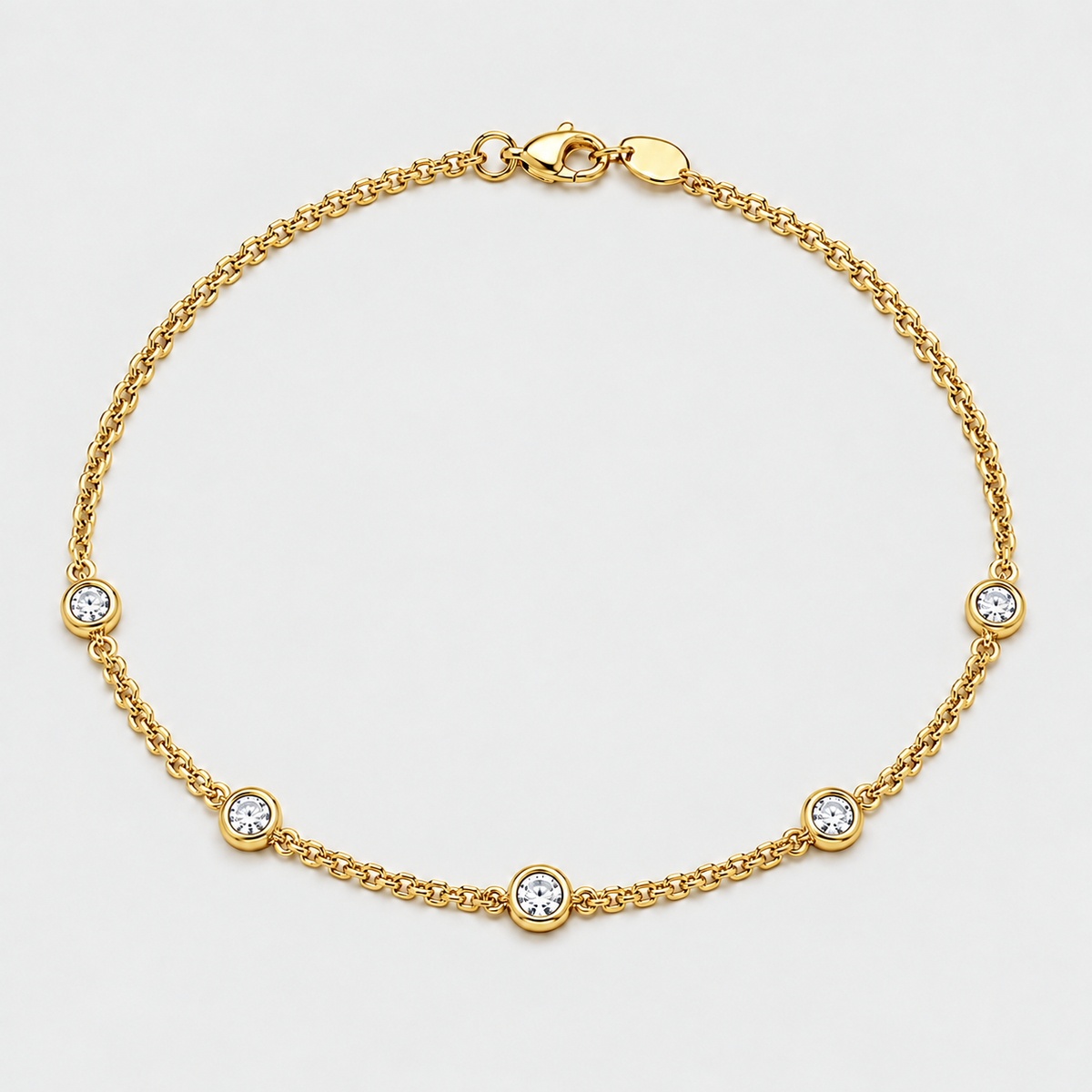 A delicate gold chain anklet featuring five evenly spaced round bezel-set clear crystals on a light gray background.