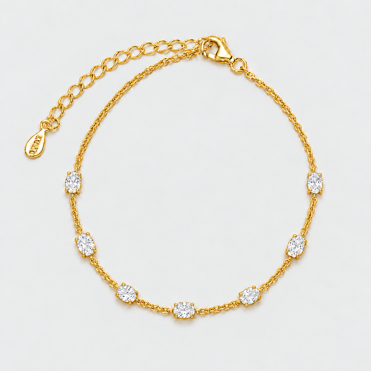 A gold chain anklet featuring seven bezel-set oval clear gemstones, displayed on a clean white background. The anklet includes an adjustable gold chain with a lobster clasp and an extension chain with a small branded tag.