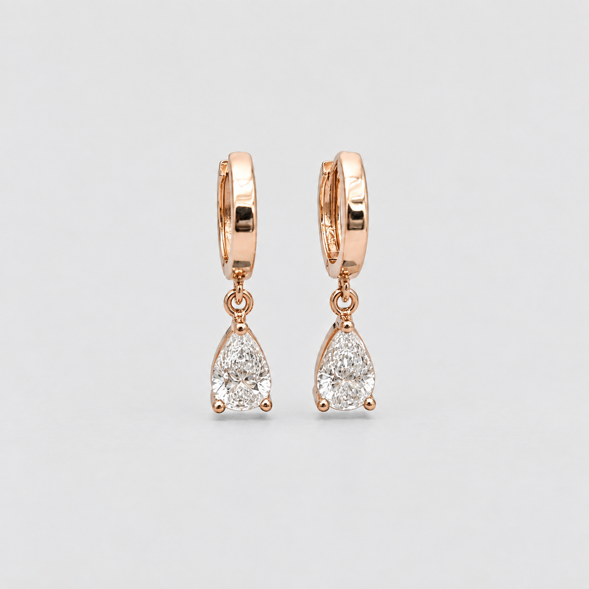 A pair of rose gold huggie hoop earrings with teardrop-shaped clear gemstones, displayed on a light gray background.