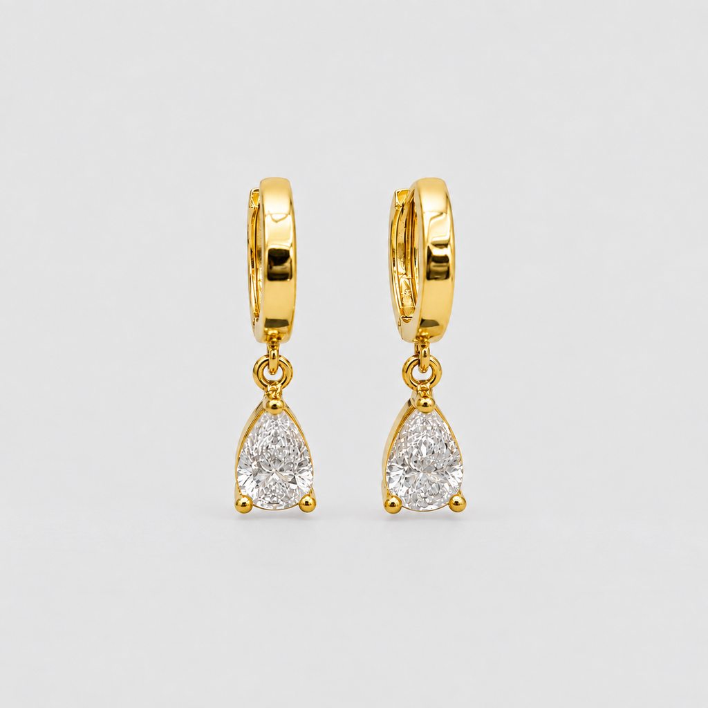 A pair of gold huggie hoop earrings with dangling pear-cut clear gemstones, shown on a light gray background.