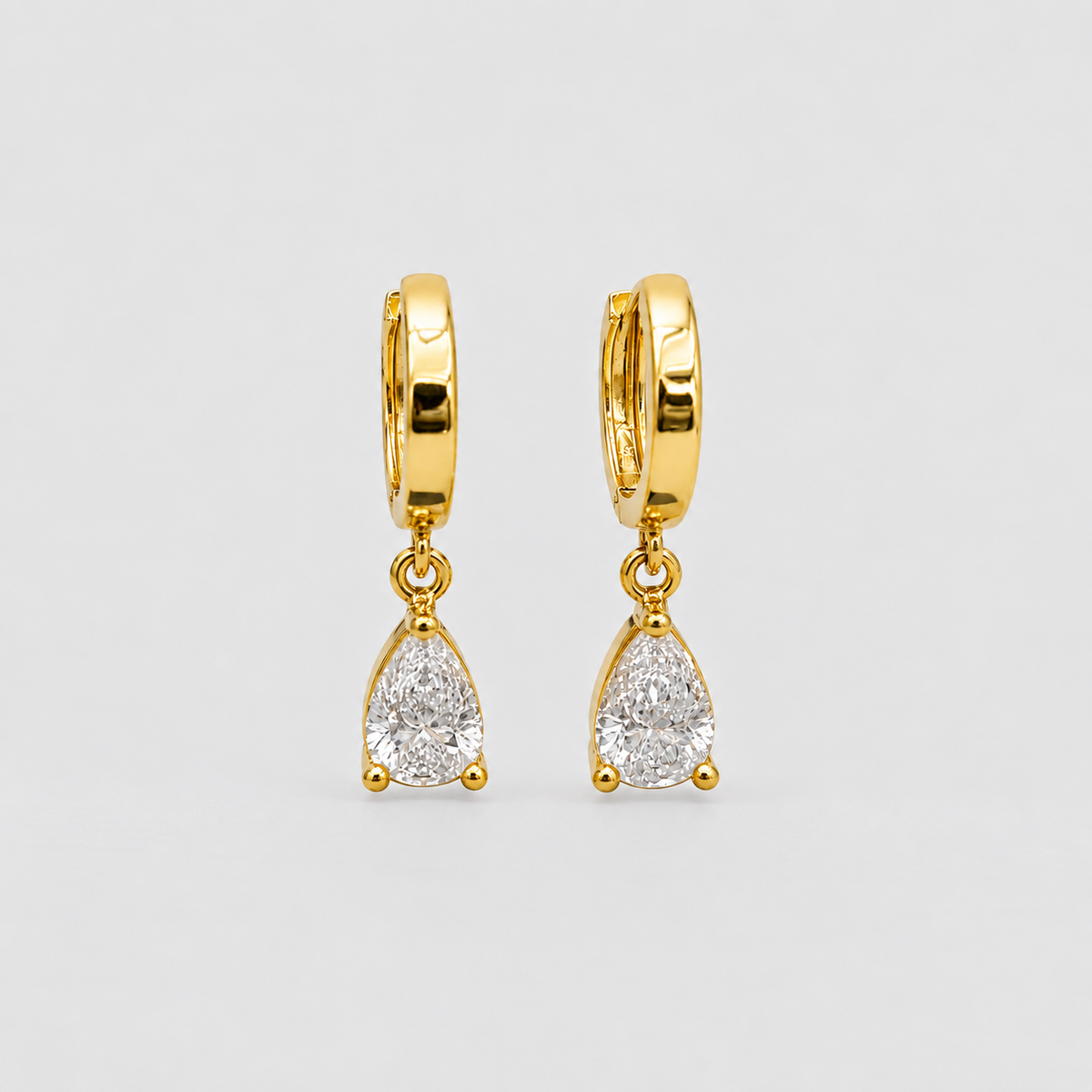 A pair of gold huggie hoop earrings with dangling pear-cut clear gemstones, shown on a light gray background.