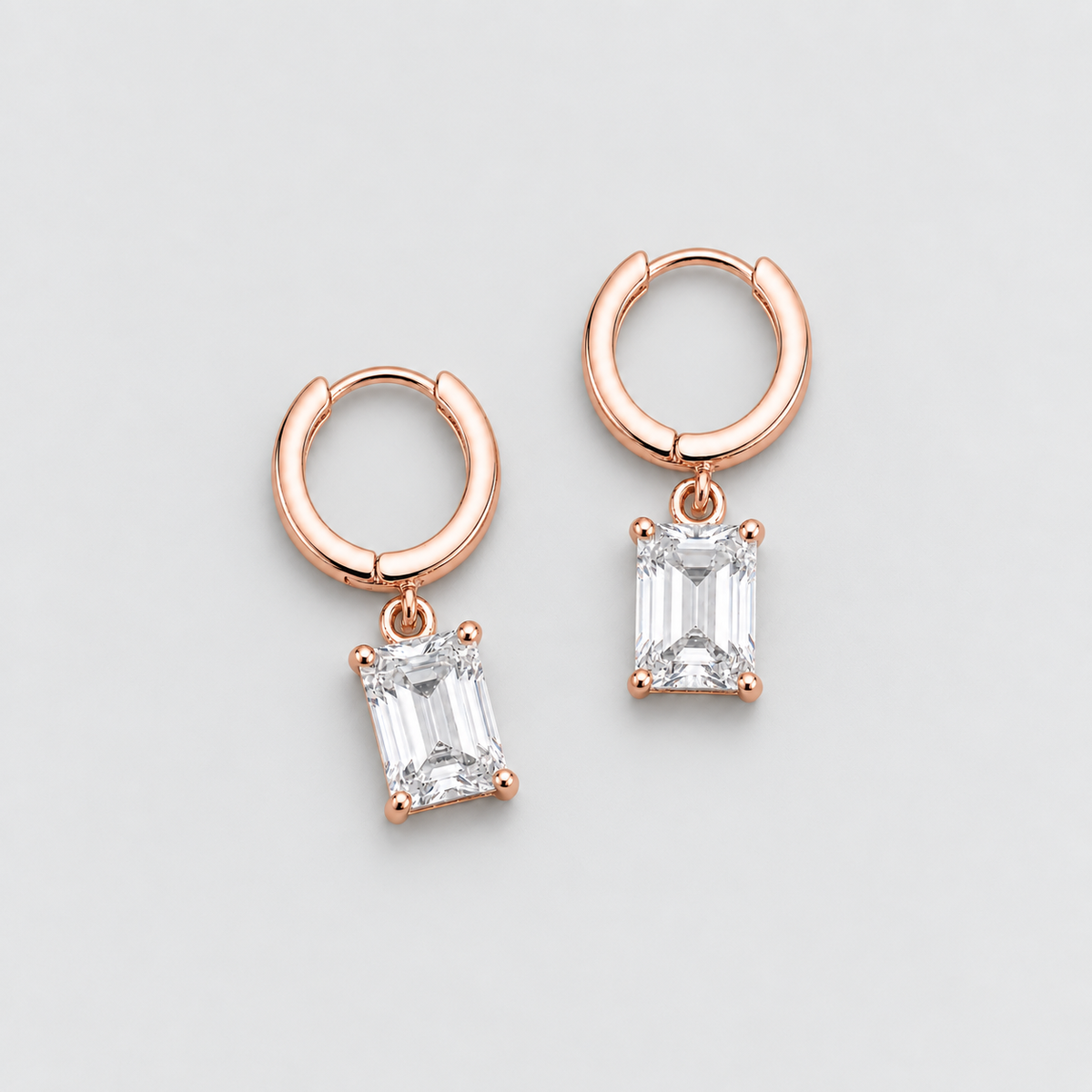 A pair of rose gold huggie hoop earrings with rectangular emerald-cut clear gemstones, displayed on a light gray background.