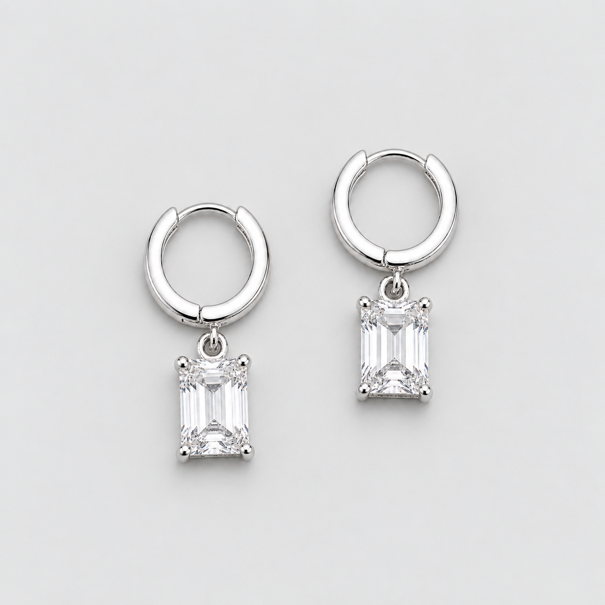 A pair of silver huggie hoop earrings with rectangular emerald-cut clear gemstones, displayed on a clean light gray background.