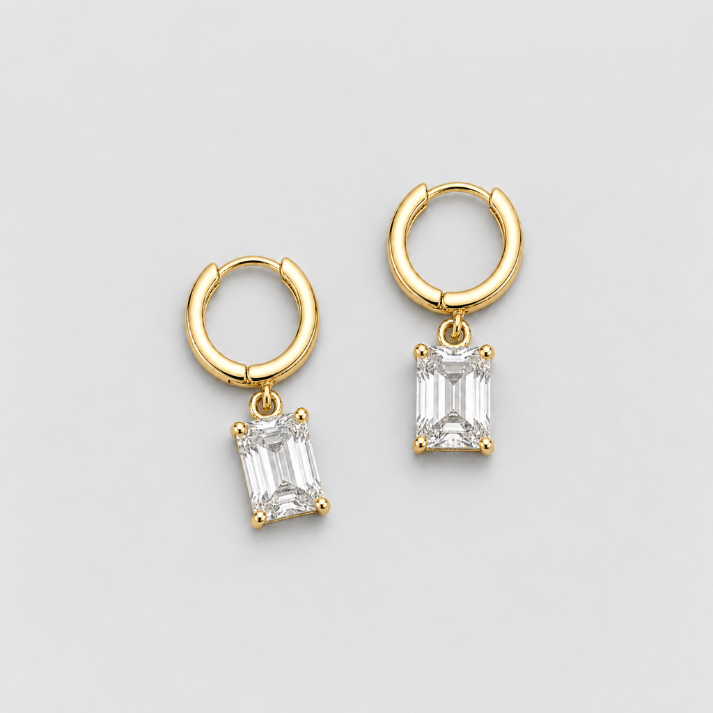 A pair of gold huggie hoop earrings with rectangular emerald-cut clear gemstones, displayed on a light gray background.