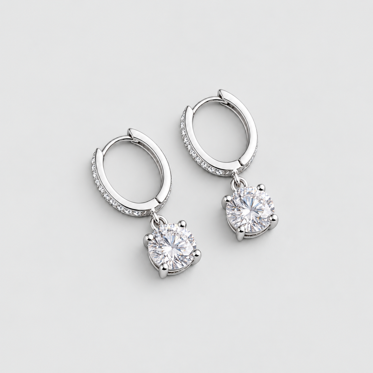 A pair of silver huggie hoop earrings with small pavé-set round stones along the front and a larger round center stone dangling below each hoop, shown on a light gray background.