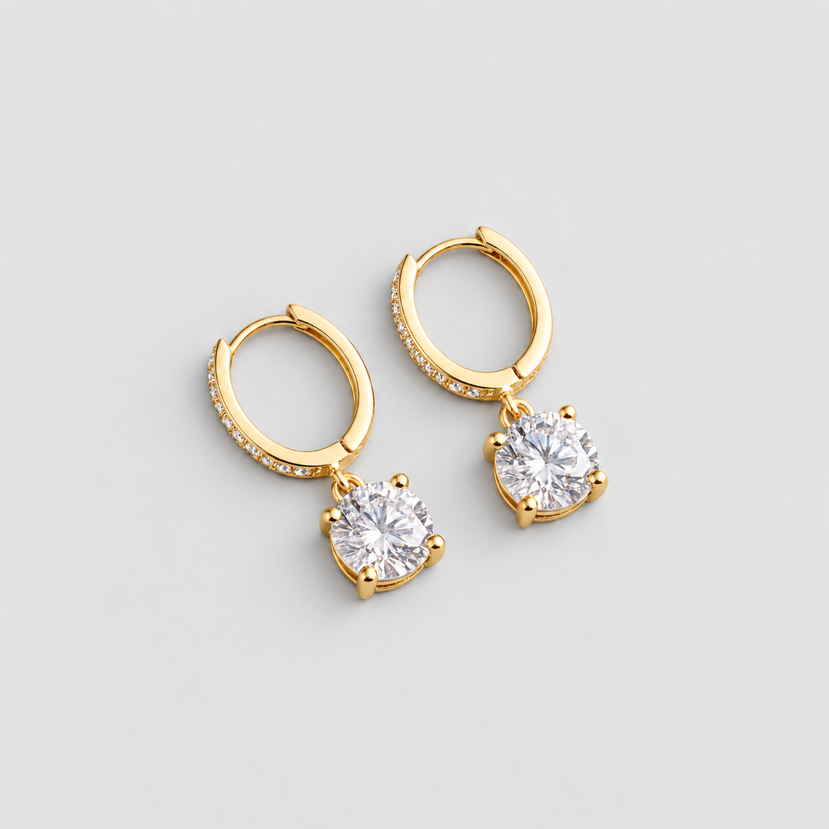 A pair of gold huggie hoop earrings with a dangling round clear gemstone in a prong setting, accented with small pavé stones along the front band, shown on a light gray background.