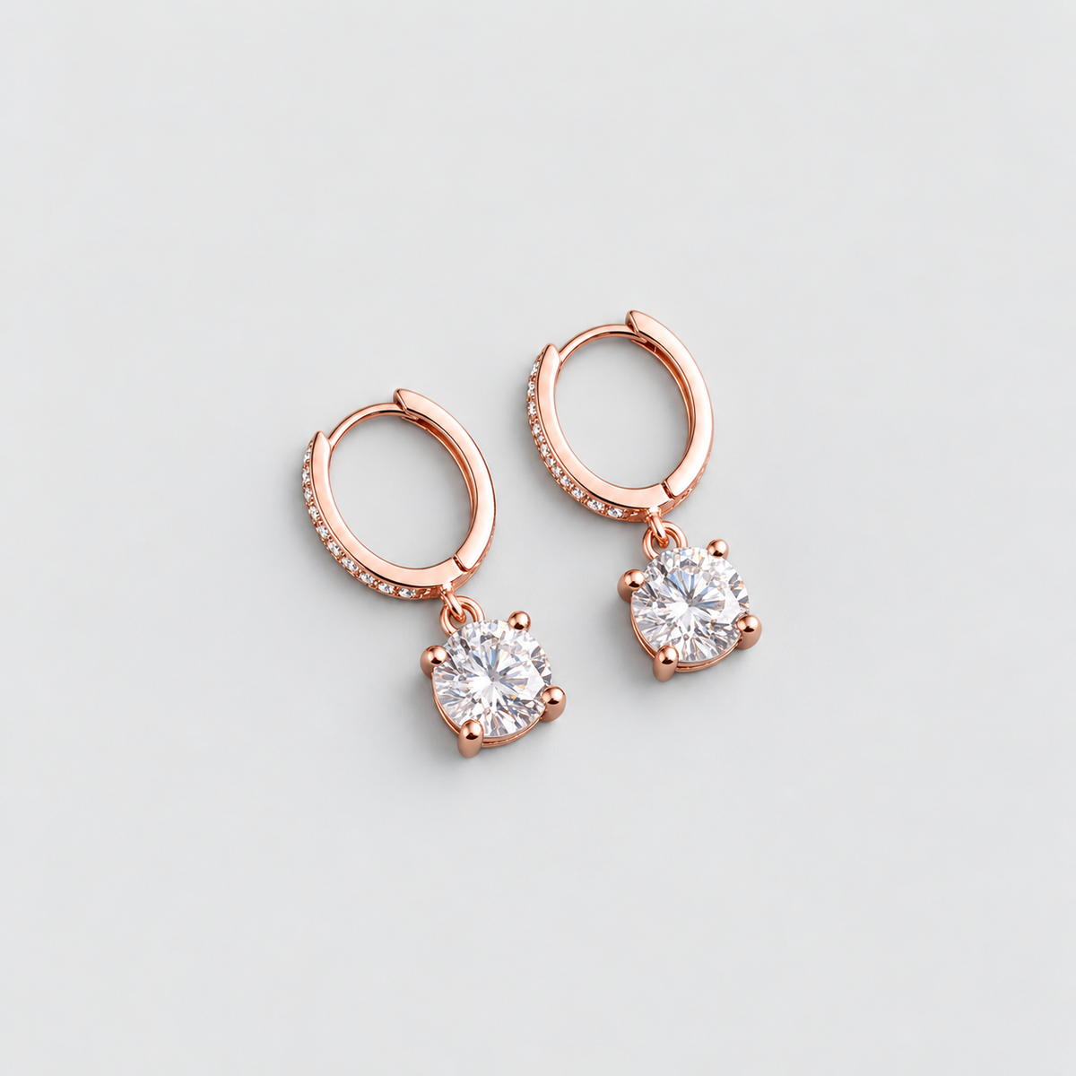 A pair of rose gold huggie hoop earrings with a dangling round-cut clear gemstone in a prong setting, shown on a light gray background.