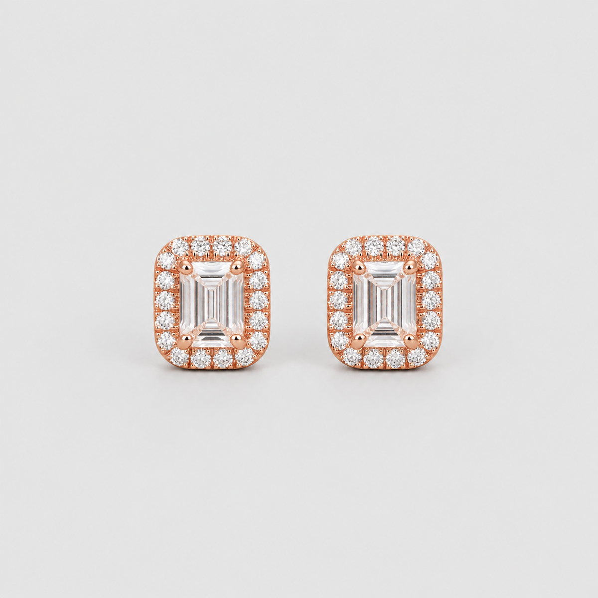 A pair of rose gold stud earrings featuring rectangular emerald-cut center stones surrounded by a halo of small clear stones, set against a light gray background.