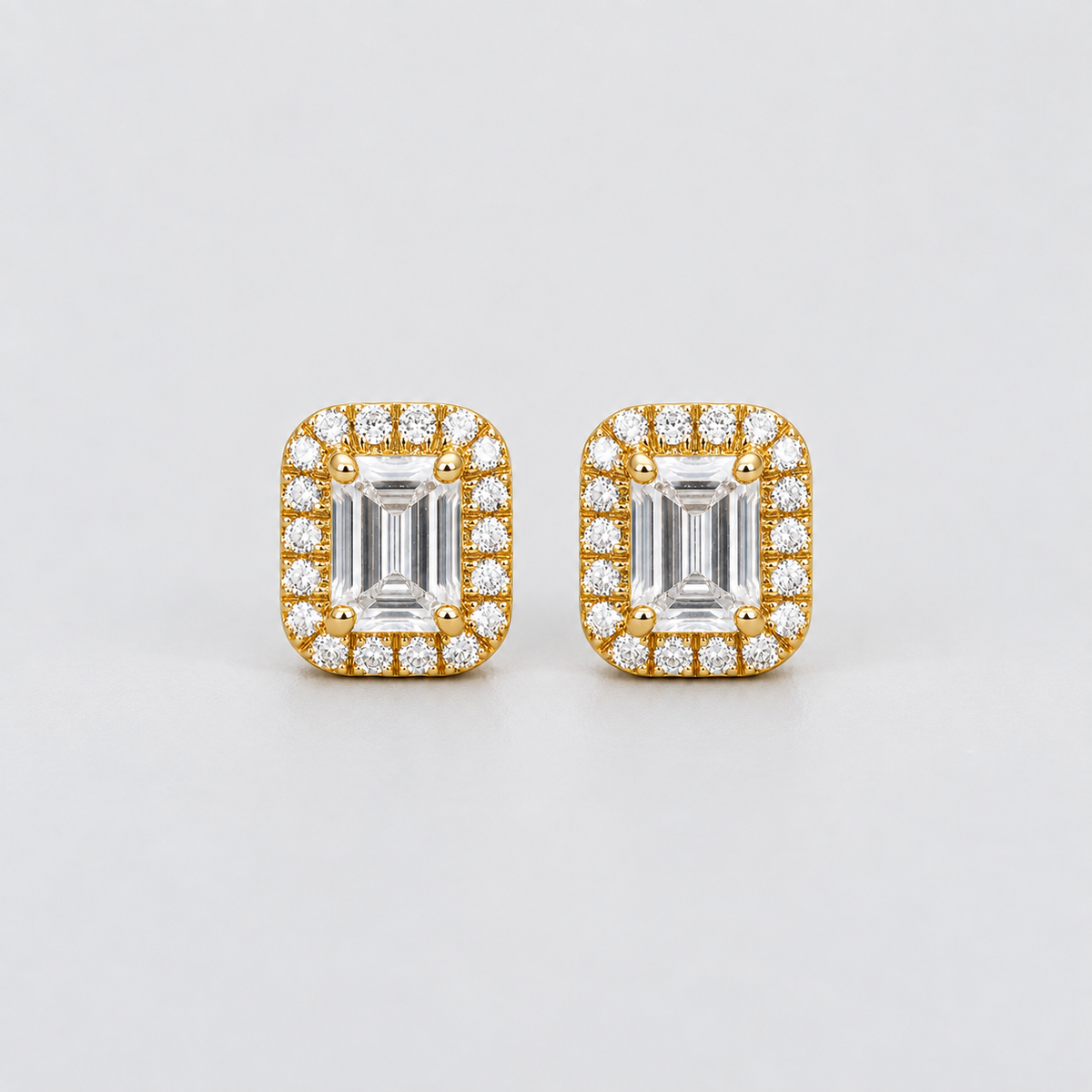 A pair of rectangular yellow gold stud earrings featuring emerald-cut center stones surrounded by a halo of small clear stones, photographed on a clean light gray background.