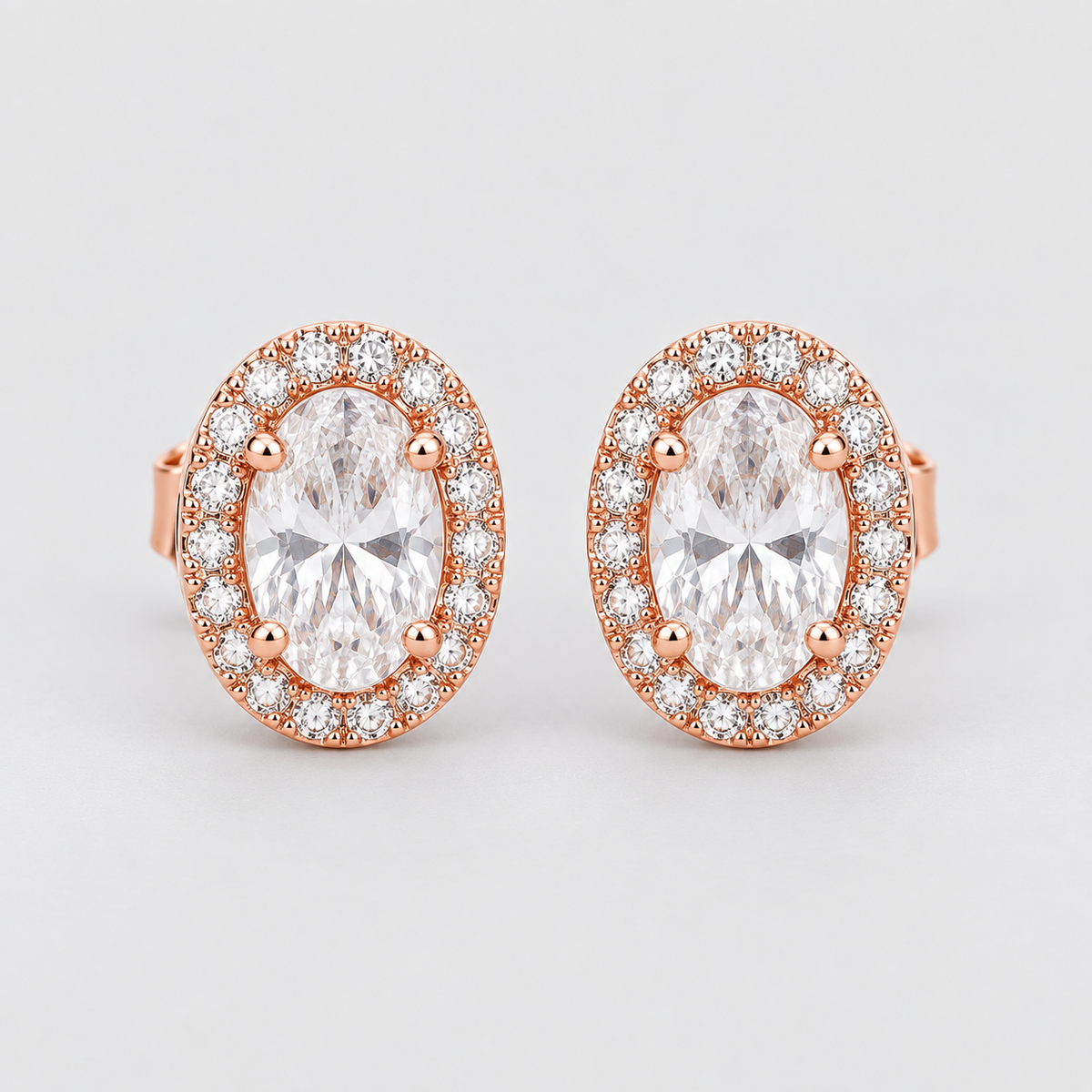 A pair of rose gold halo stud earrings featuring oval-cut clear center stones surrounded by a halo of small round clear stones, set against a light gray background.