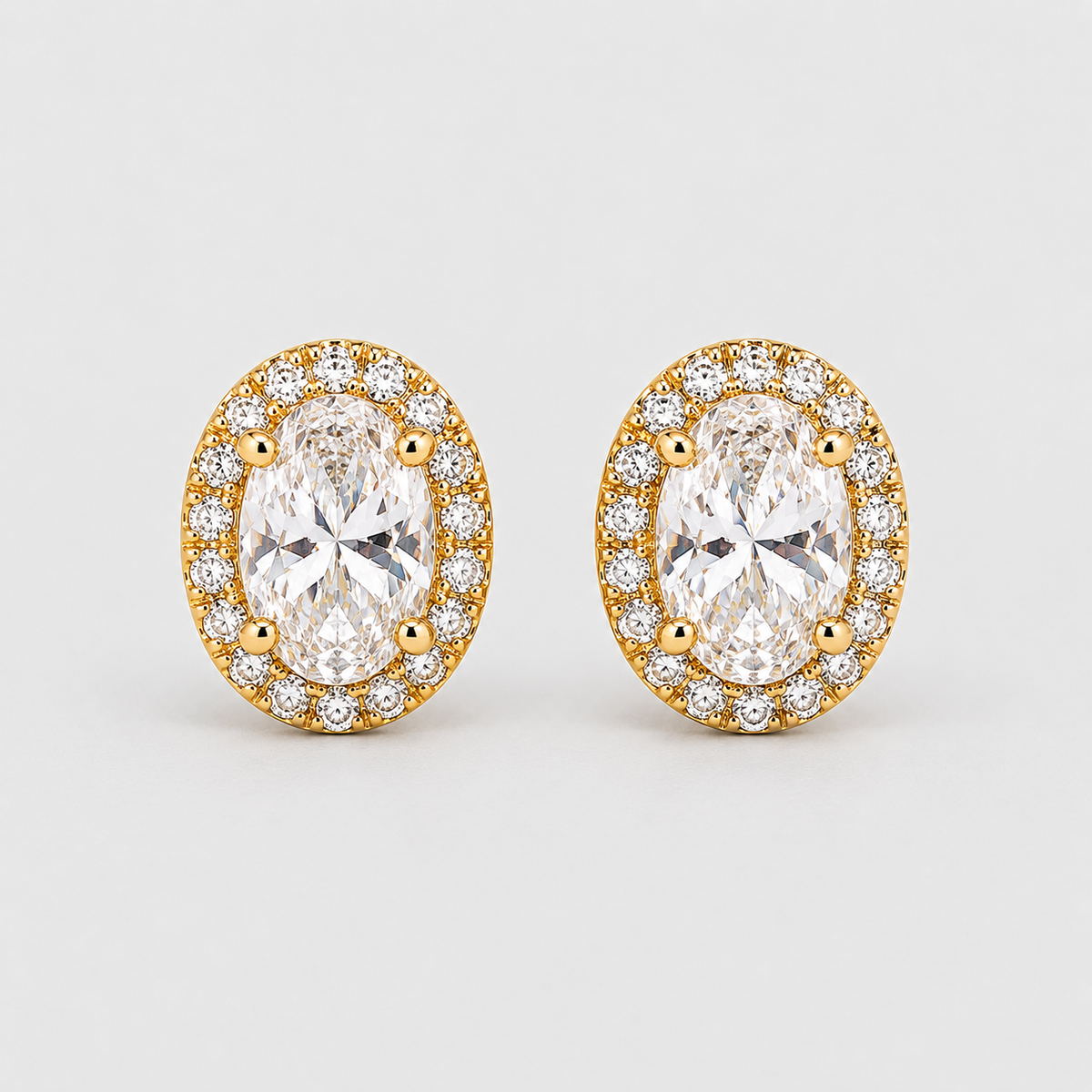 A pair of oval stud earrings featuring large clear marquise-cut center stones surrounded by a halo of smaller round clear stones in yellow gold settings, photographed on a light gray background.