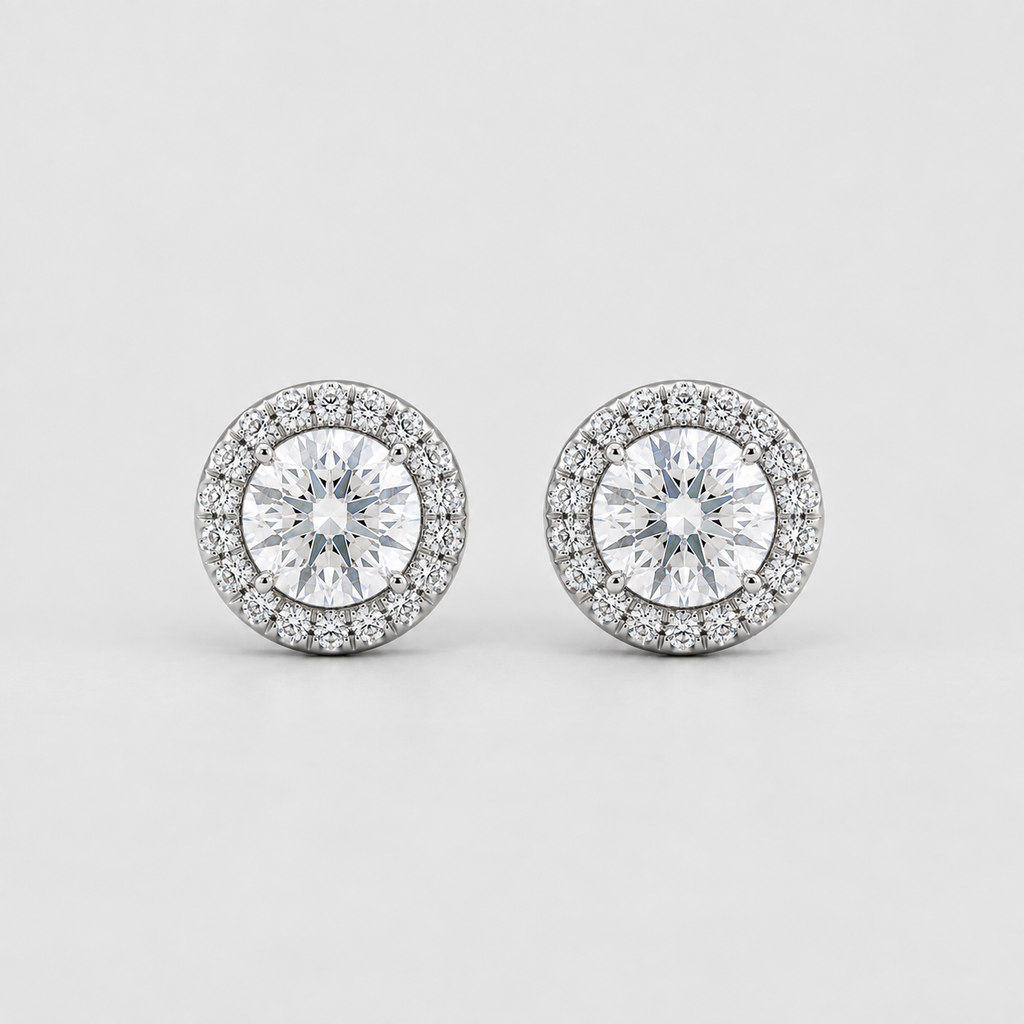 A pair of round diamond halo stud earrings in silver-toned metal, featuring a central round diamond surrounded by a halo of smaller diamonds, set against a clean light gray background.