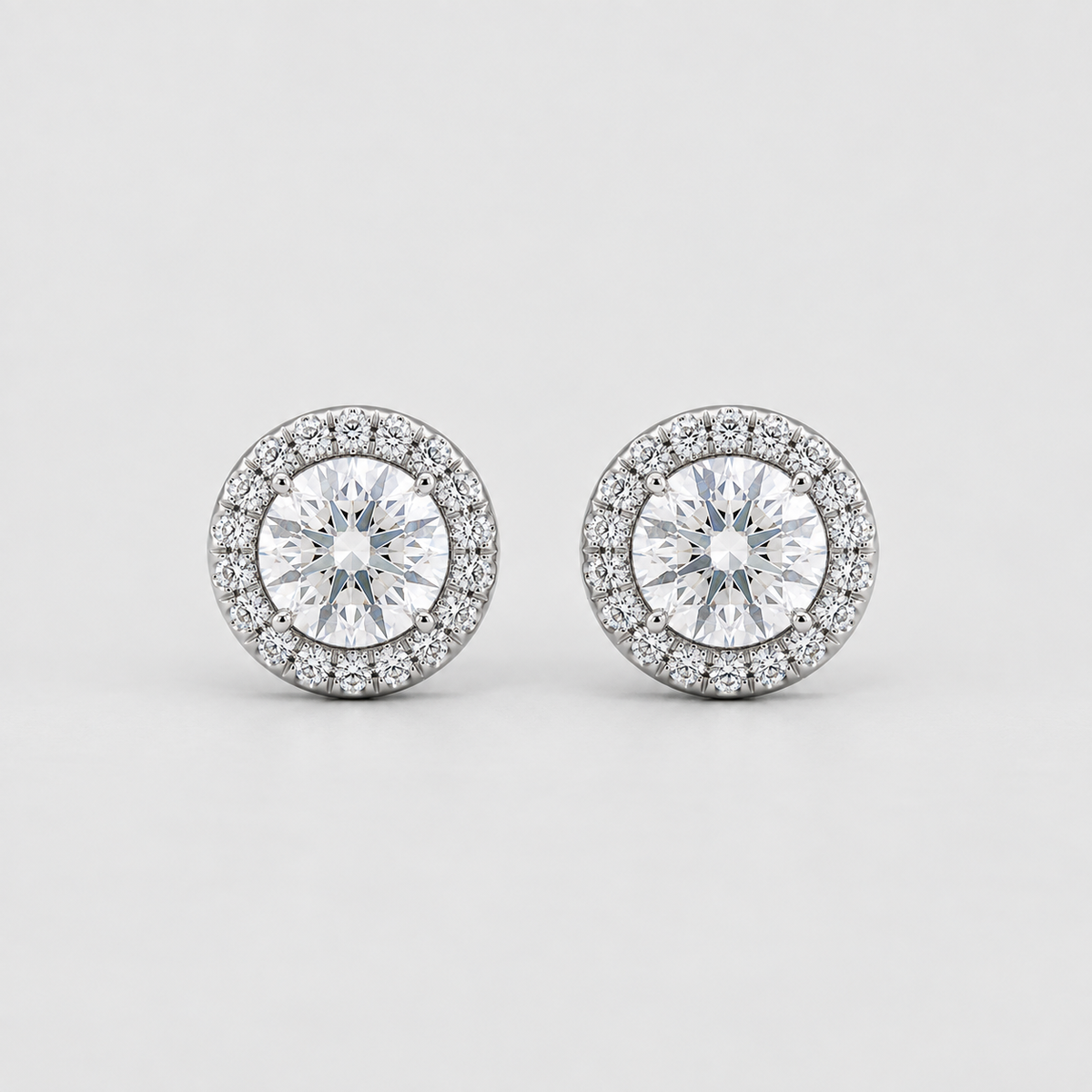 A pair of round diamond halo stud earrings in silver-toned metal, featuring a central round diamond surrounded by a halo of smaller diamonds, set against a clean light gray background.