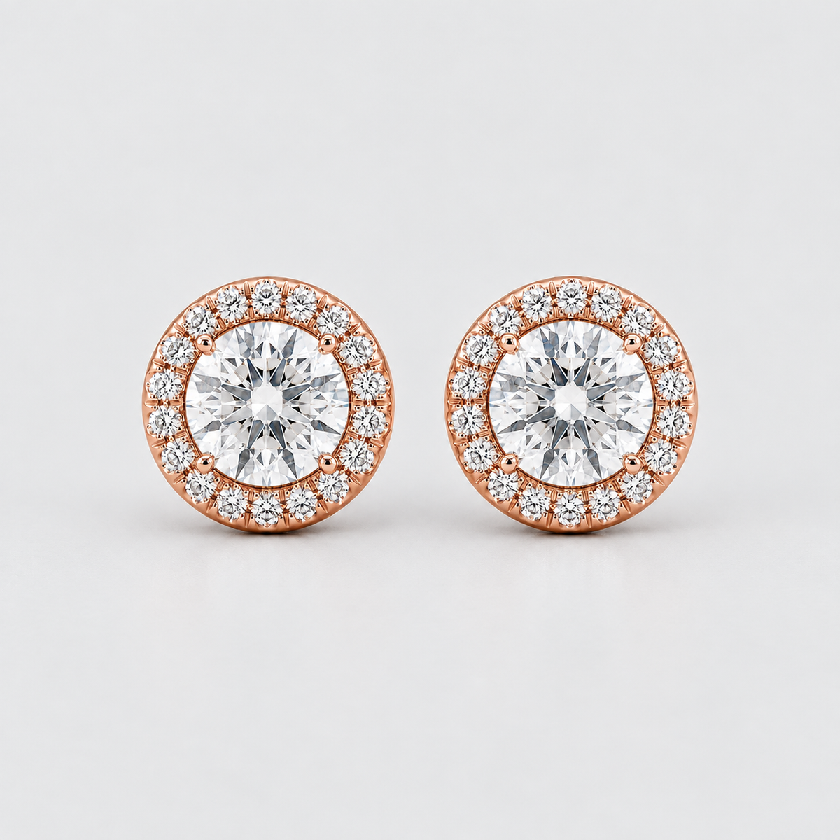 A pair of round halo stud earrings featuring a central round brilliant-cut clear gemstone surrounded by a halo of smaller clear stones in a rose gold setting, shown on a light gray background.