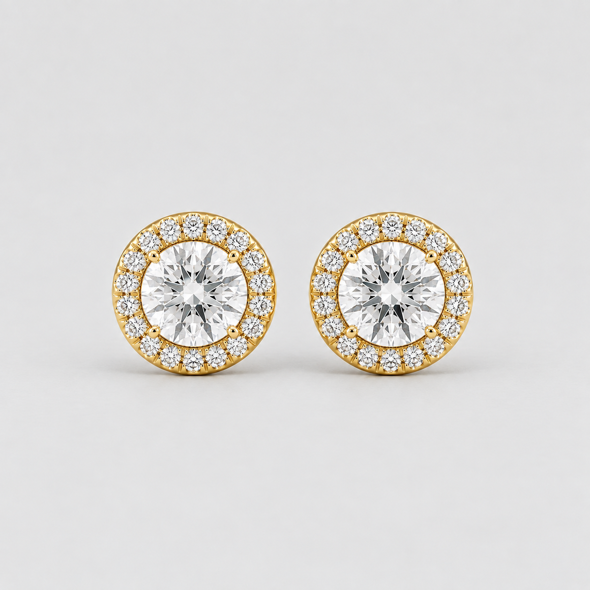 A pair of round yellow gold stud earrings, each featuring a large central clear round-cut gemstone surrounded by a halo of smaller clear pavé-set gemstones, photographed on a light gray background.