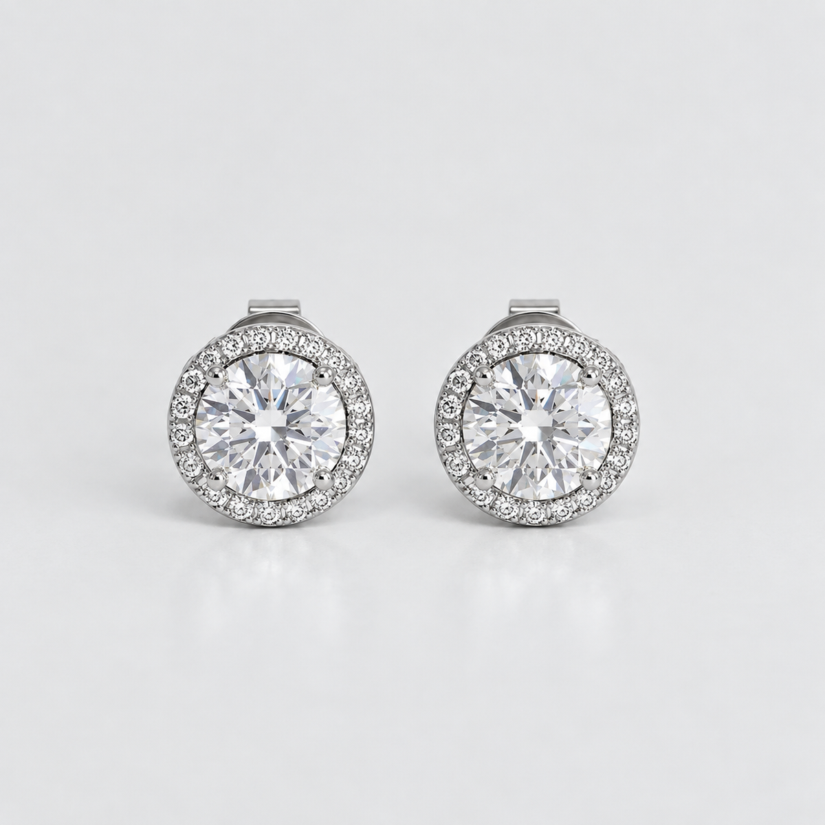A pair of round halo stud earrings with large clear center stones and a border of smaller clear stones in a silver-toned setting, photographed on a clean light gray background.