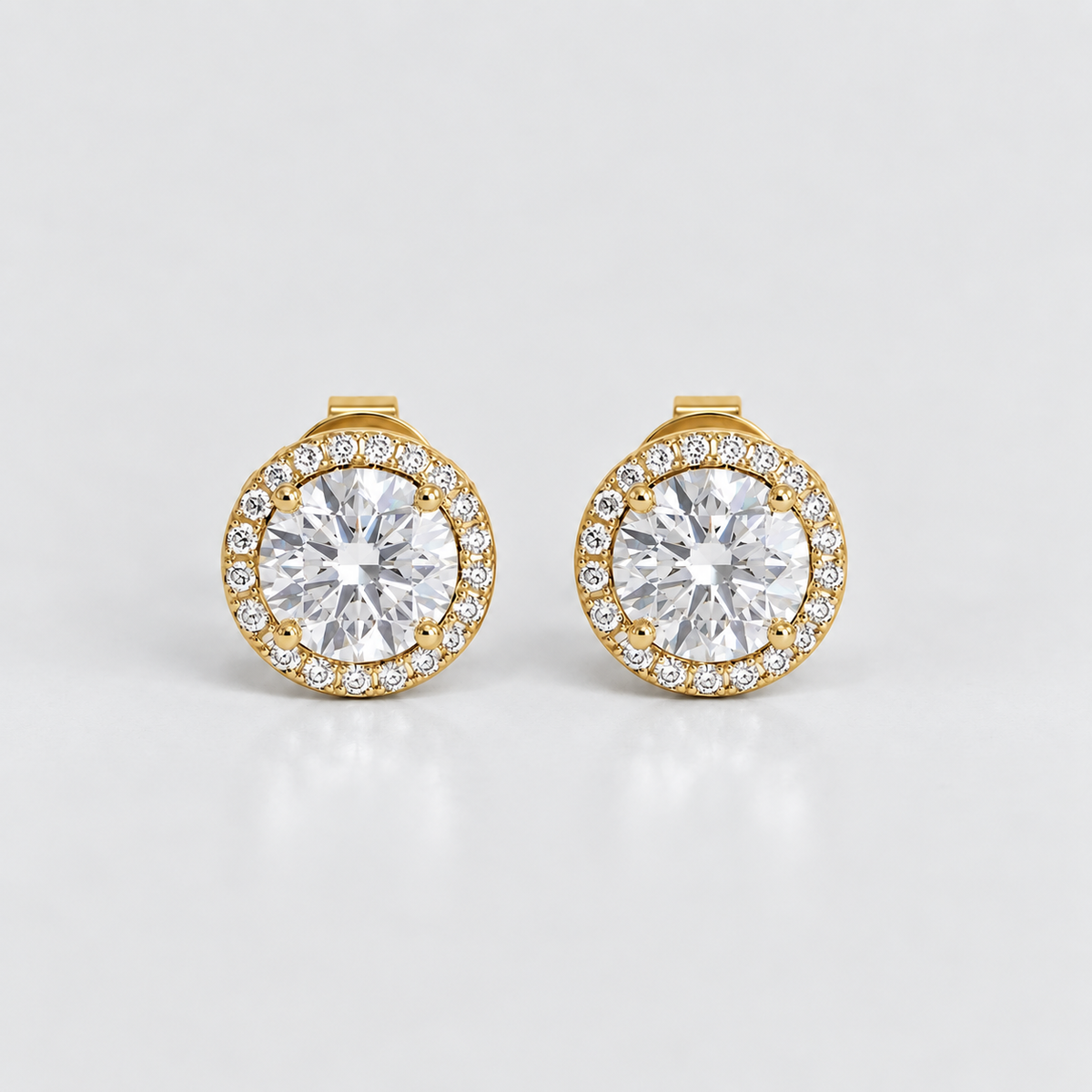 A pair of gold round stud earrings with a halo of small clear stones surrounding a larger central clear gemstone, shown on a light gray reflective background.