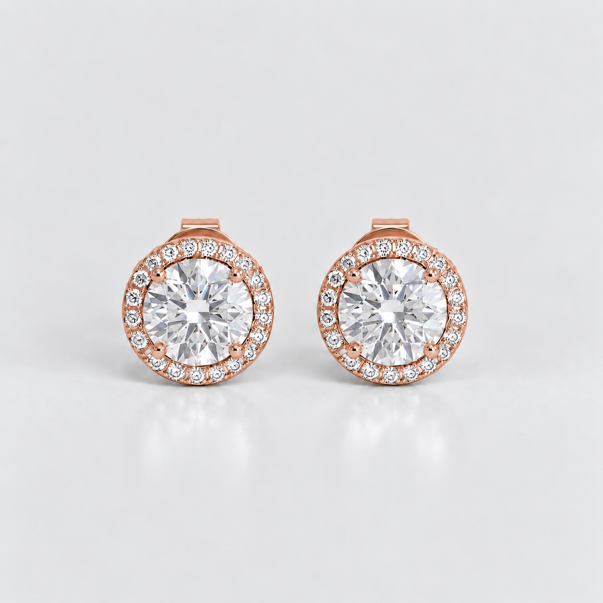 A pair of rose gold stud earrings with a halo setting, featuring a large round clear center stone surrounded by a border of smaller round stones, set against a light gray background.