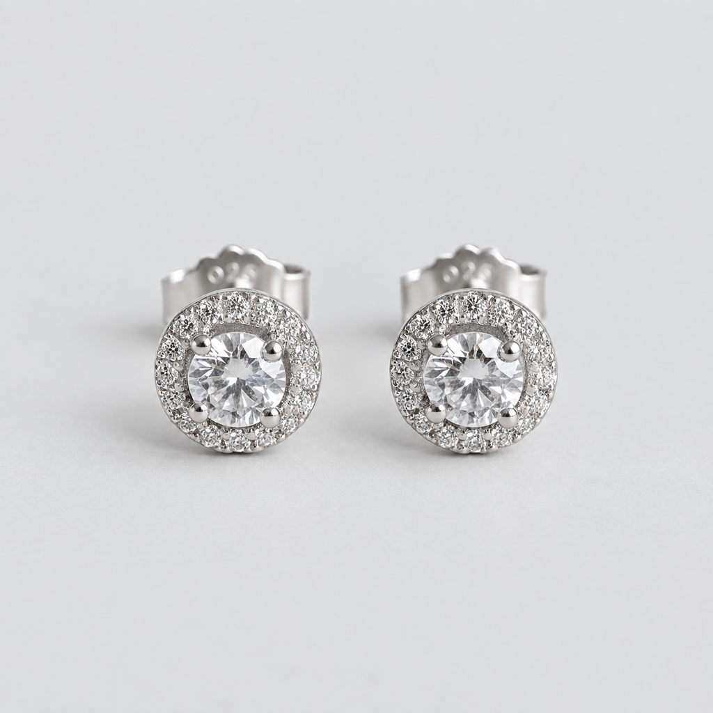 A pair of round halo stud earrings with large clear center stones and a surrounding halo of smaller clear stones in a silver-toned setting, photographed on a light gray background.