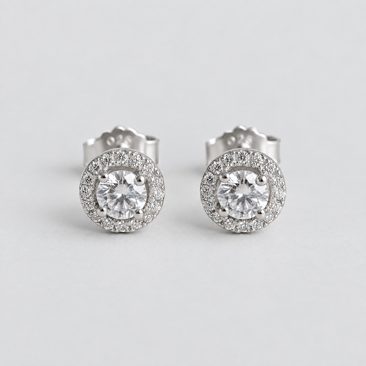 A pair of round halo stud earrings with large clear center stones and a surrounding halo of smaller clear stones in a silver-toned setting, photographed on a light gray background.