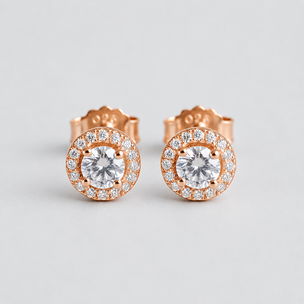 A pair of rose gold stud earrings with round-cut clear center stones surrounded by a halo of smaller clear stones, shown on a light gray background.