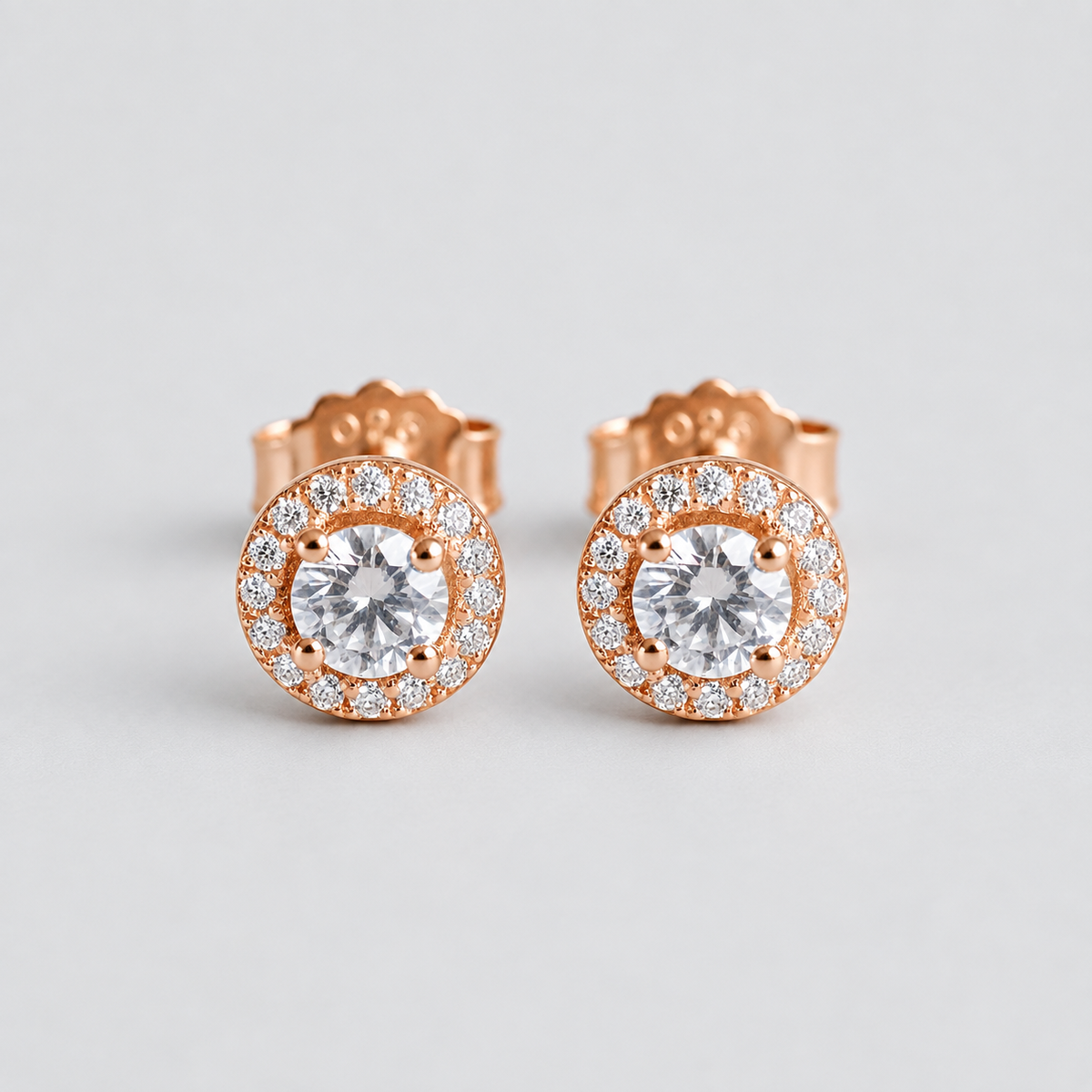 A pair of rose gold stud earrings with round-cut clear center stones surrounded by a halo of smaller clear stones, shown on a light gray background.