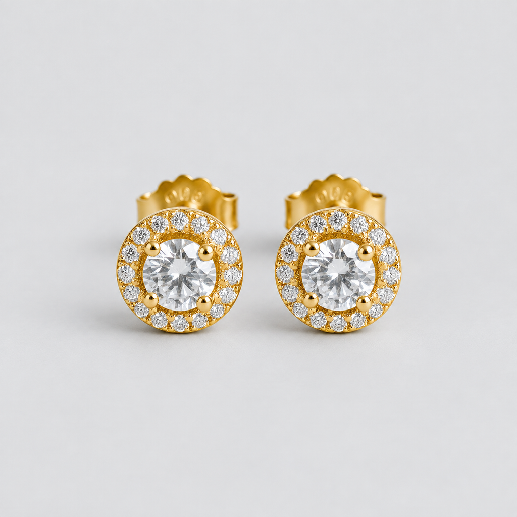 A pair of gold stud earrings with round-cut clear center stones surrounded by a halo of smaller clear stones, photographed on a clean light gray background.