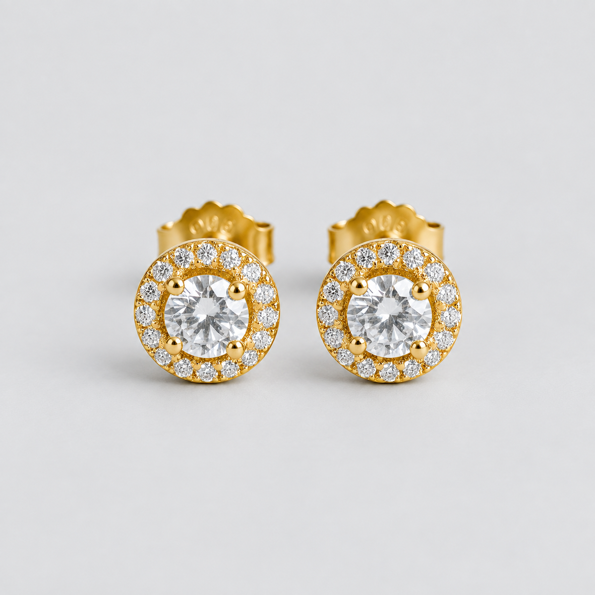 A pair of gold stud earrings with round-cut clear center stones surrounded by a halo of smaller clear stones, photographed on a clean light gray background.