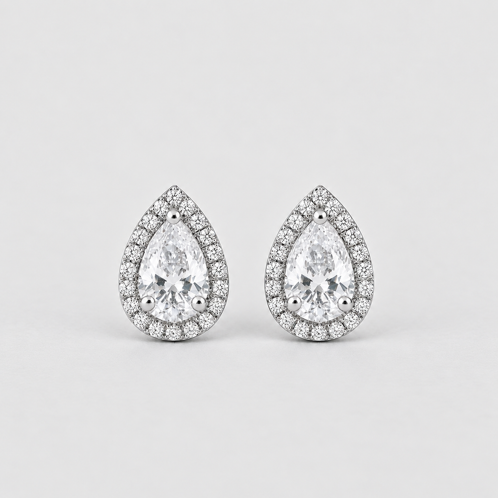A pair of pear-shaped halo stud earrings featuring a large clear center stone surrounded by a border of smaller round stones in a silver-toned setting, shown on a light gray background.