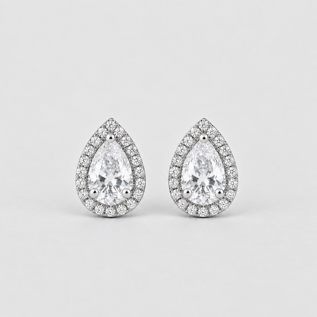 A pair of pear-shaped halo stud earrings featuring a large clear center stone surrounded by a border of smaller round stones in a silver-toned setting, shown on a light gray background.