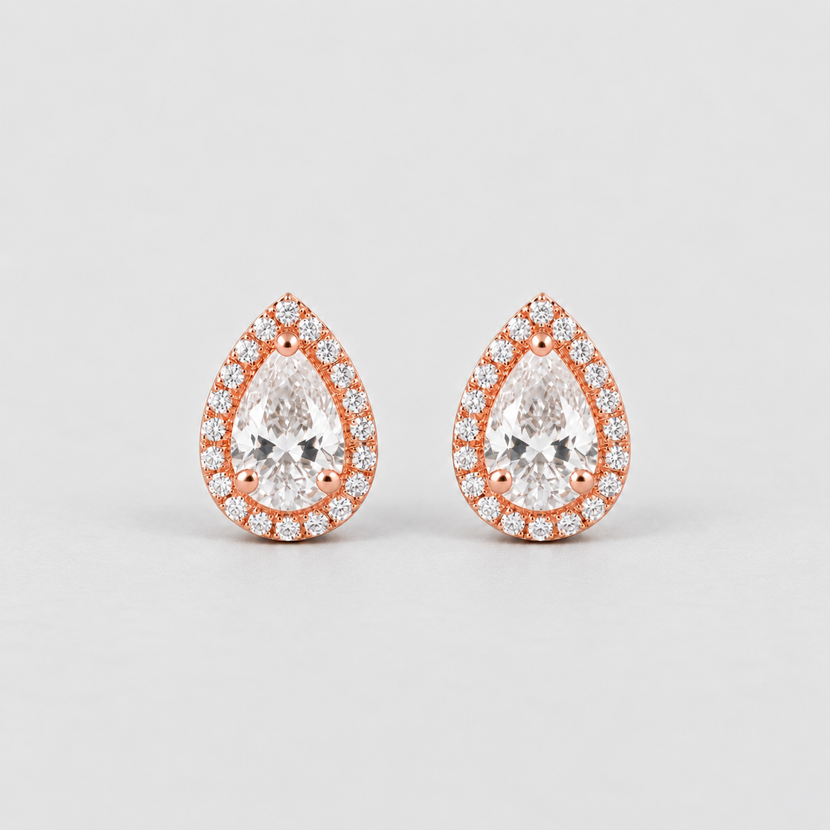 A pair of pear-shaped stud earrings in rose gold with a halo of small clear stones around a larger central clear stone, shown on a light gray background.