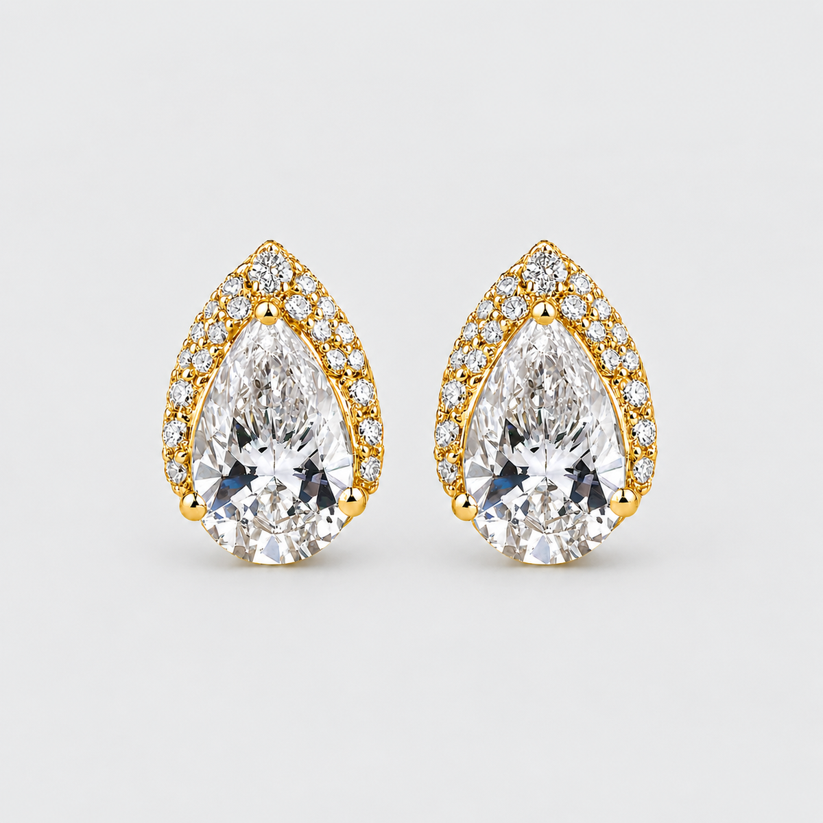 A pair of pear-shaped gold stud earrings with large clear center stones and smaller clear stones along the border, shown on a light gray background.