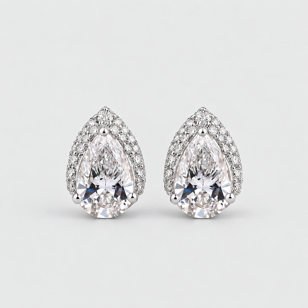 A pair of pear-shaped diamond stud earrings in a silver-toned setting, surrounded by small round diamonds, on a light gray background.