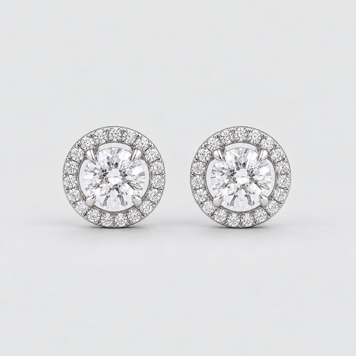 A pair of round halo stud earrings featuring a central round-cut clear gemstone surrounded by a halo of smaller clear stones in a silver-toned setting, shown on a light gray background.
