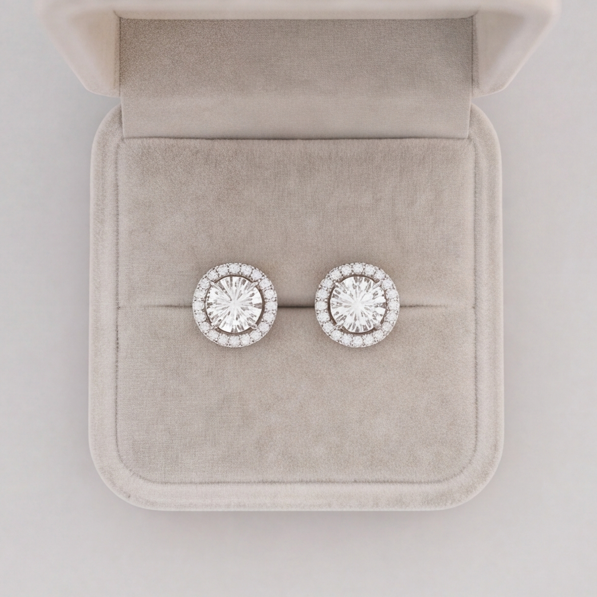A pair of round halo stud earrings with clear gemstones in a silver-toned setting, displayed on a light beige velvet jewelry box.