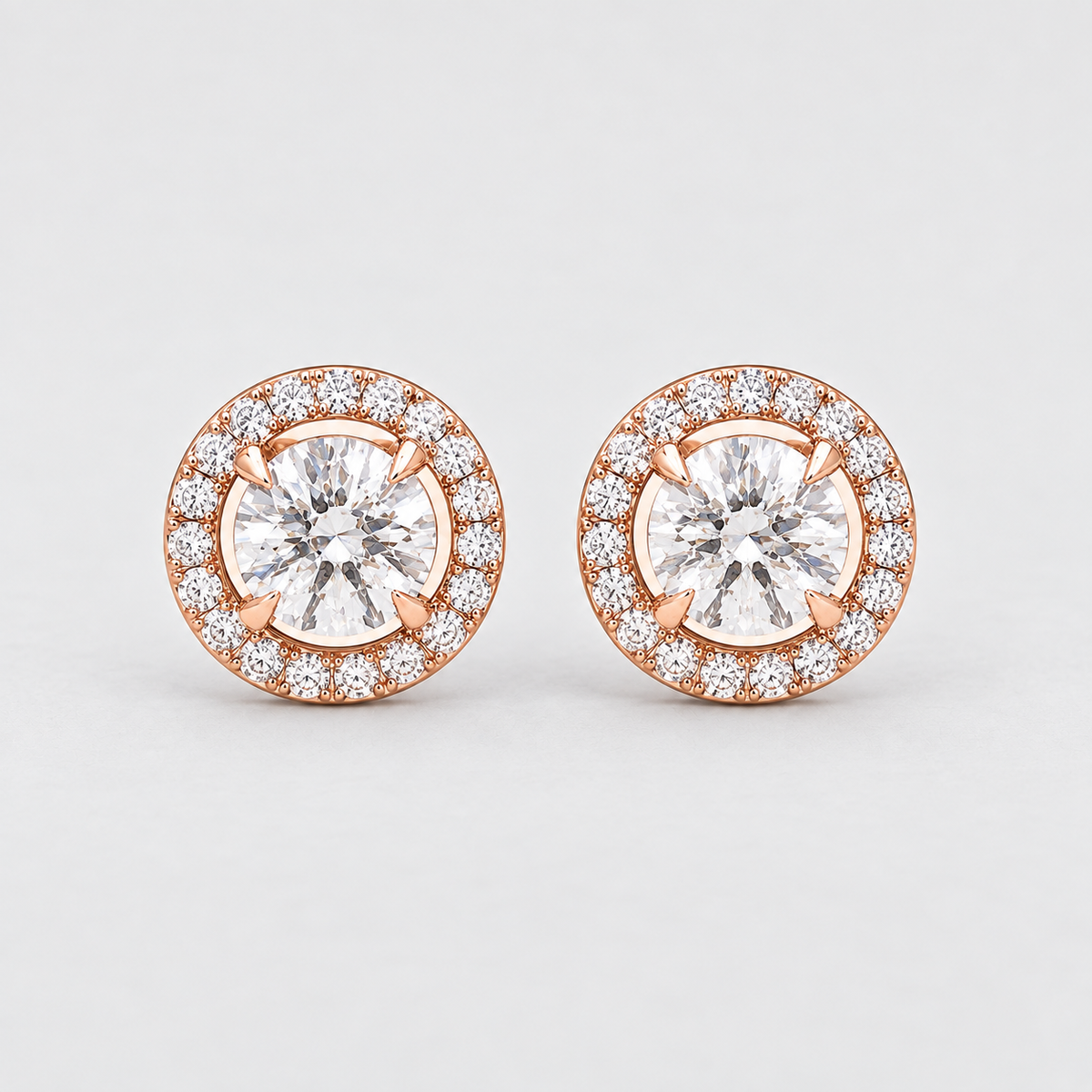 A pair of round halo stud earrings featuring a central clear gemstone surrounded by a halo of smaller clear stones in a rose gold setting, photographed on a light gray background.