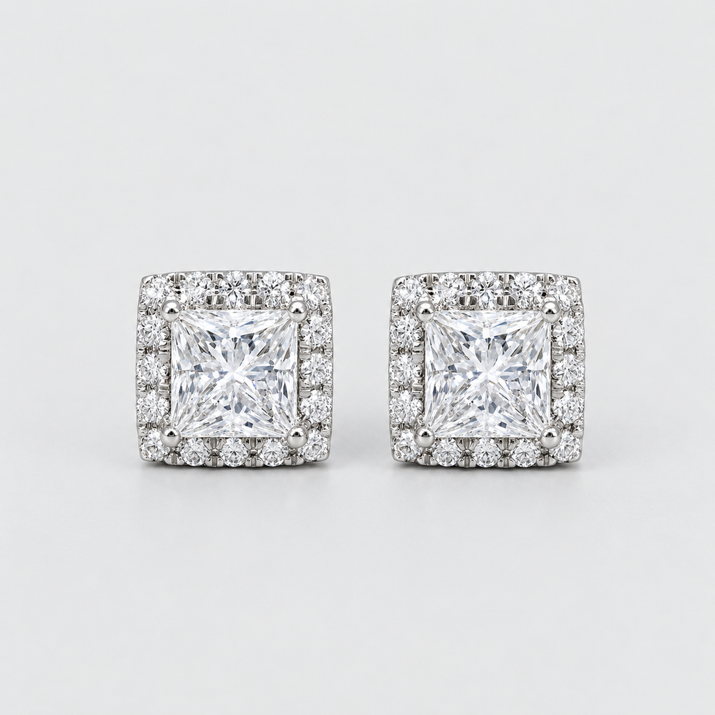 A pair of square stud earrings featuring a princess-cut clear center stone surrounded by a halo of smaller clear stones, set in a silver-toned metal on a light gray background.
