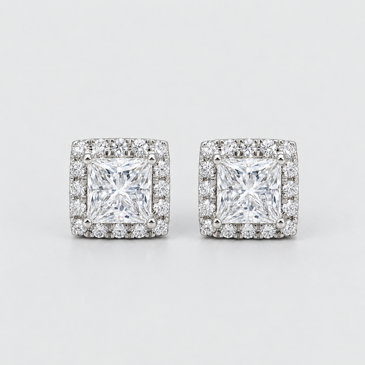 A pair of square stud earrings featuring a princess-cut clear center stone surrounded by a halo of smaller clear stones, set in a silver-toned metal on a light gray background.