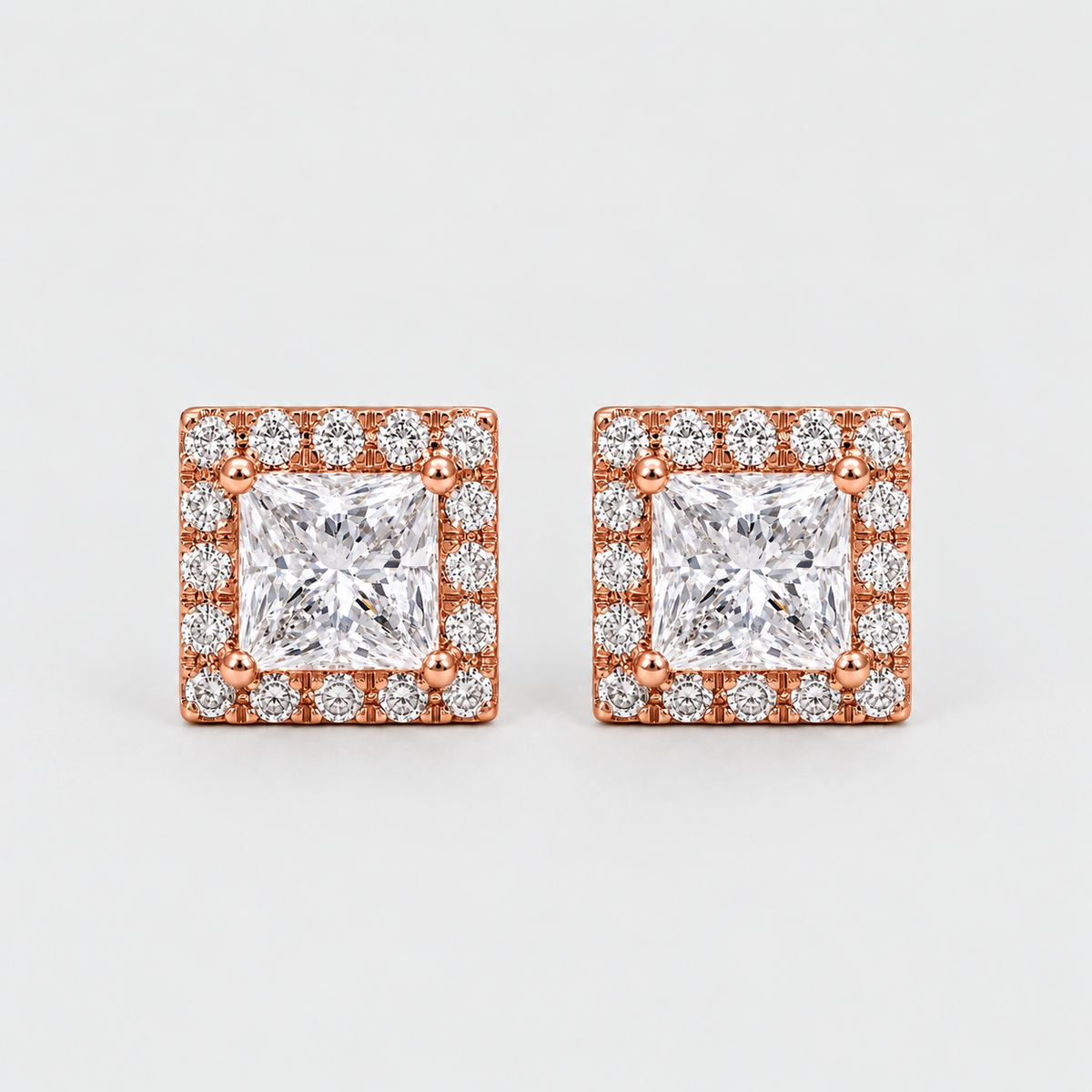 A pair of square stud earrings featuring a princess-cut clear center stone surrounded by a halo of smaller clear stones in a rose gold setting, photographed on a clean light gray background.