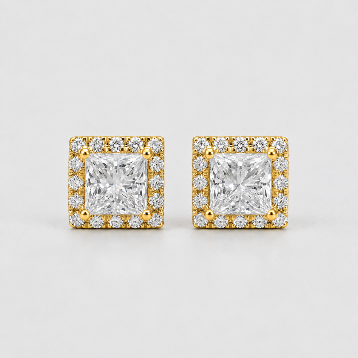 A pair of square stud earrings featuring a central princess-cut clear gemstone surrounded by a halo of smaller clear stones in a yellow gold setting, photographed on a light gray background.