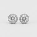 A pair of round halo stud earrings featuring large clear center stones surrounded by a halo of smaller clear stones in a silver-toned setting, shown on a light gray background.