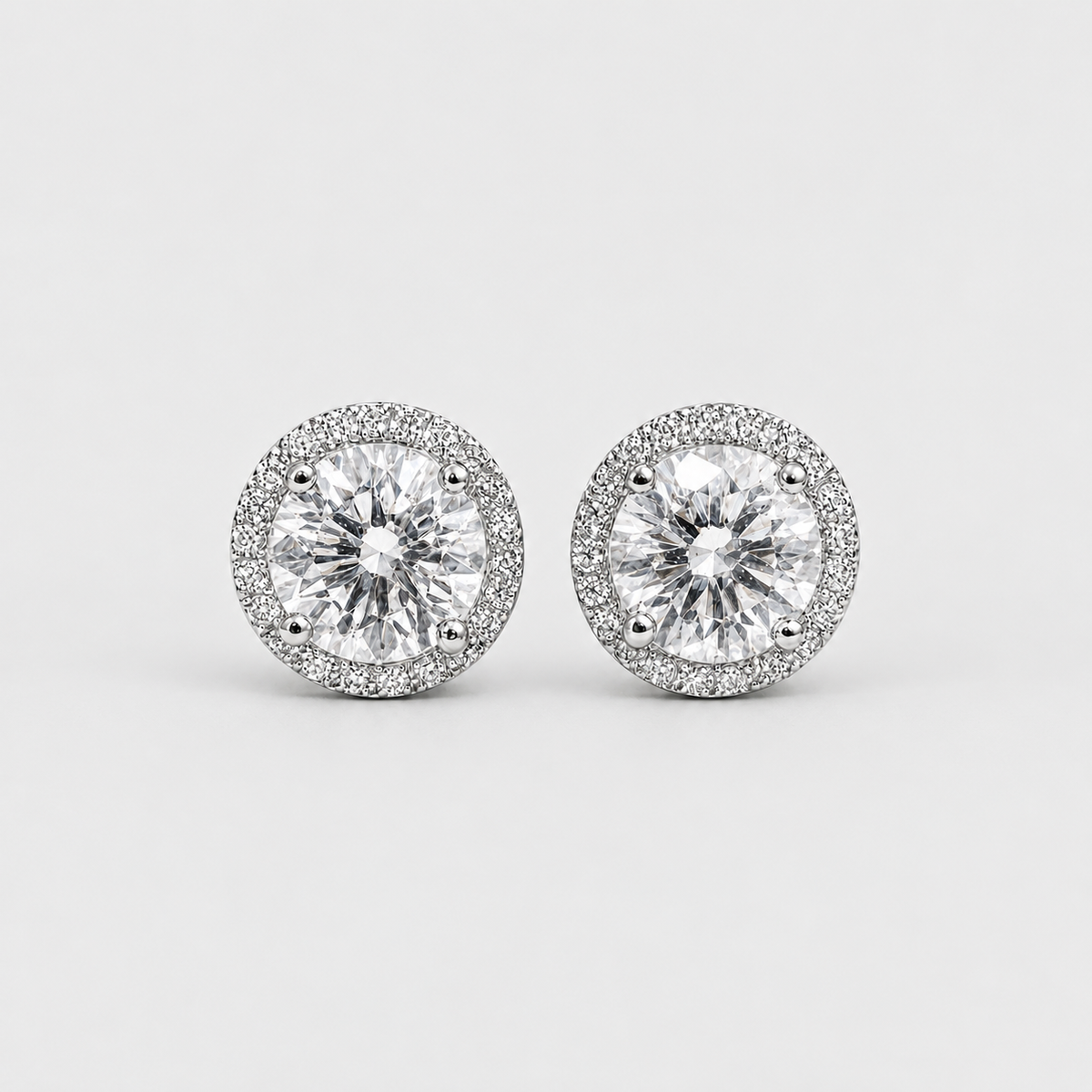 A pair of round halo stud earrings featuring large clear center stones surrounded by a halo of smaller clear stones in a silver-toned setting, shown on a light gray background.